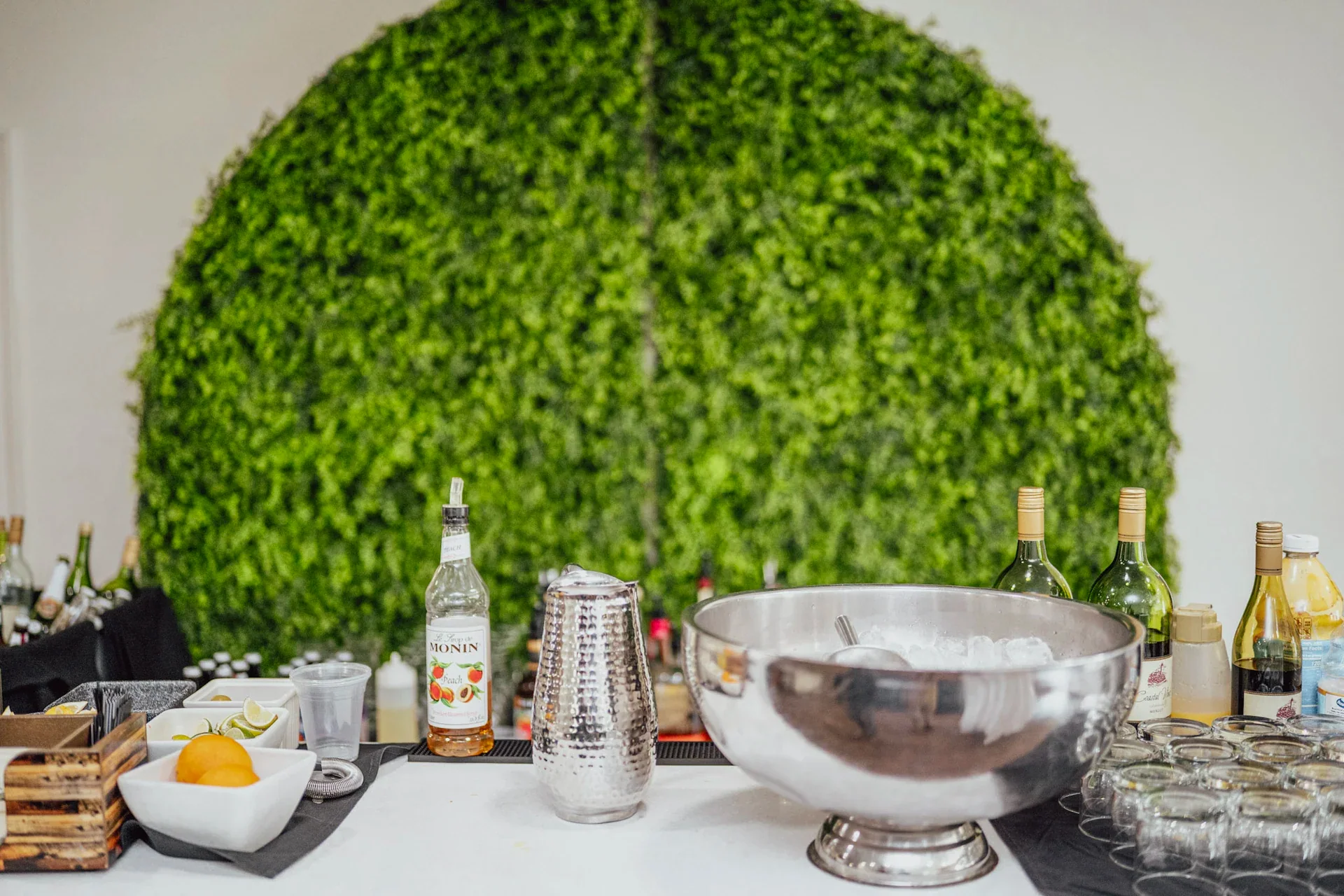 A drink station with fruit, bottles and a large metal bowl against a green wall