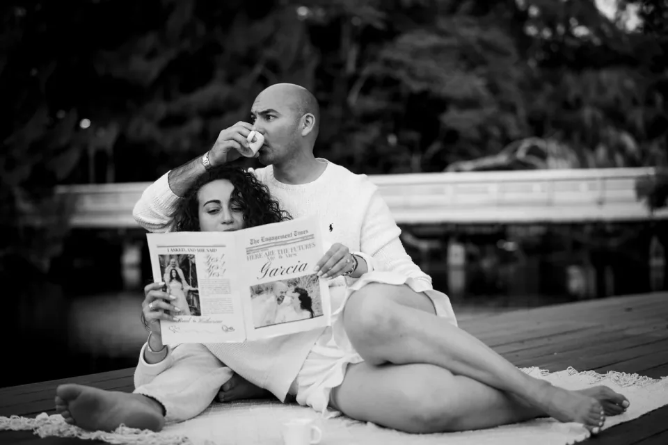 Couple reclining together with a newspaper beside a lake, in black and white