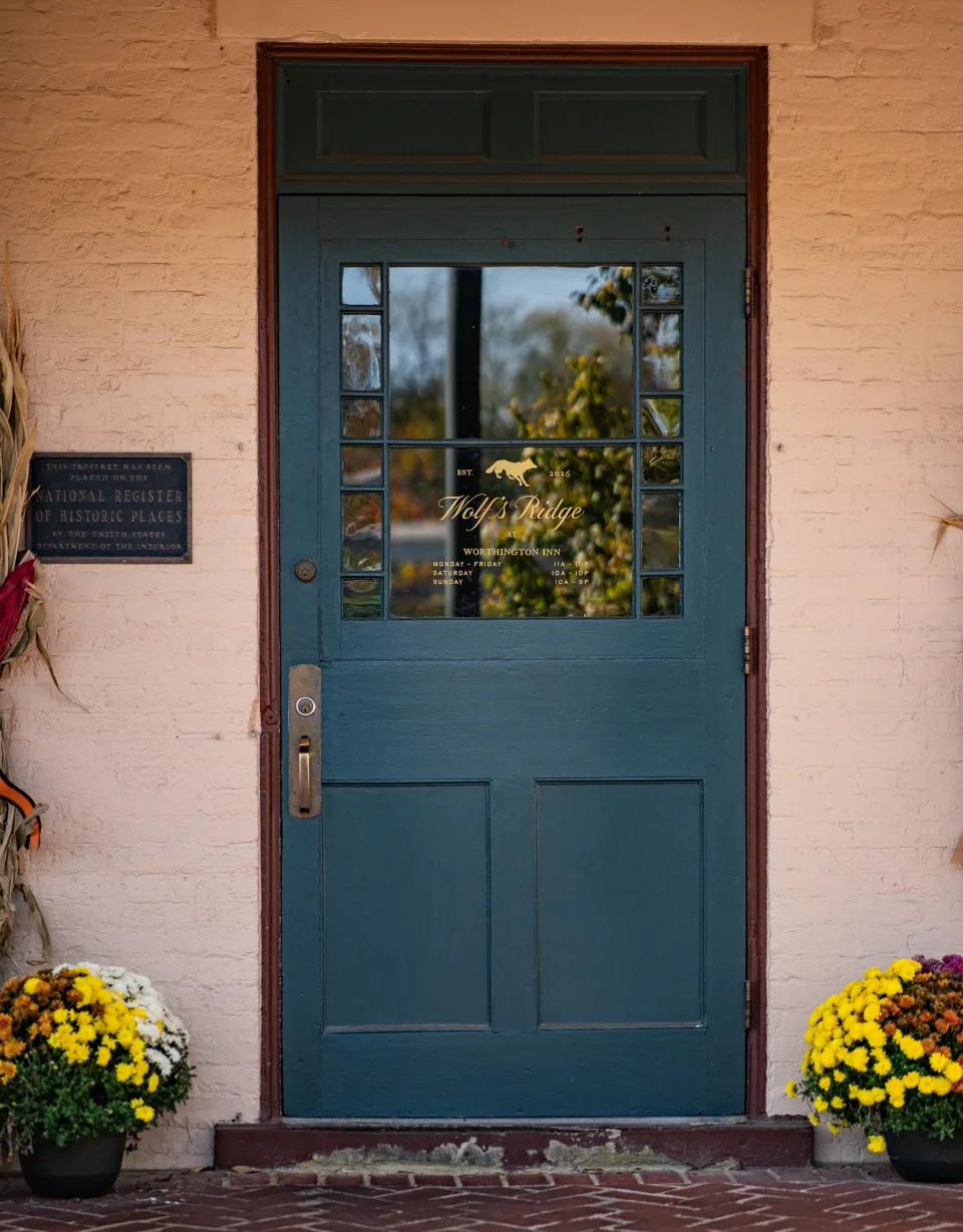 A teal shop door framed by warm brick and pots of yellow flowers