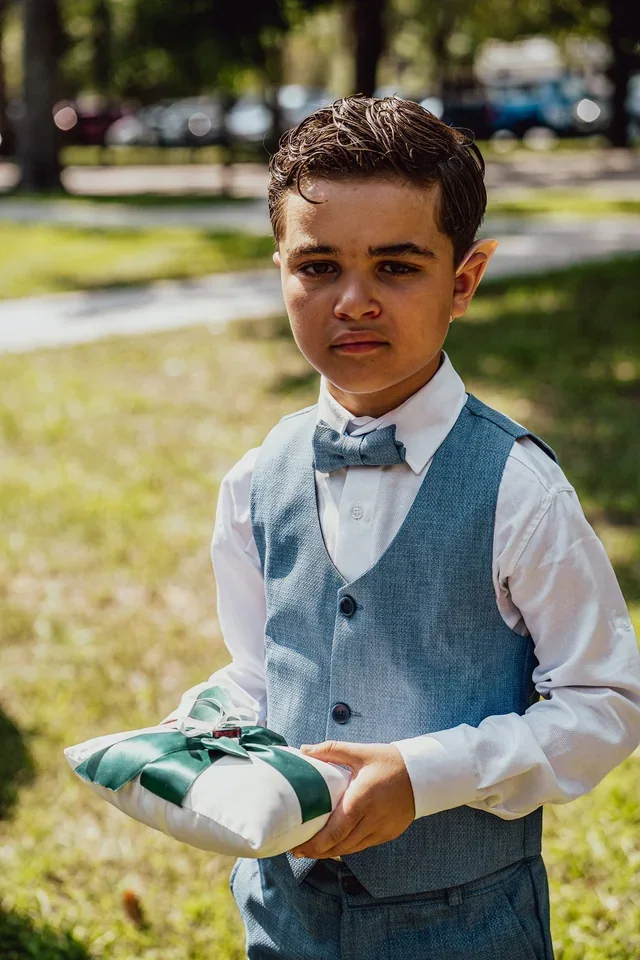 A child in a vest and bow tie holding a ring pillow outdoors