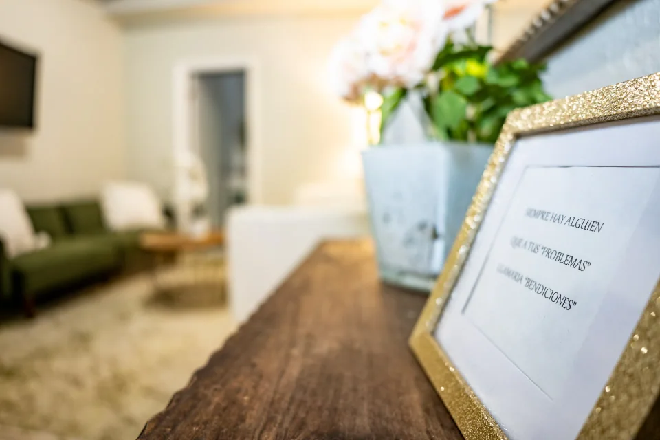 A framed sign and vase of flowers on a wooden counter with a sitting room softly blurred behind