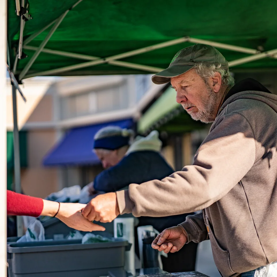 A market vendor reaching toward a customer beneath a green canopy