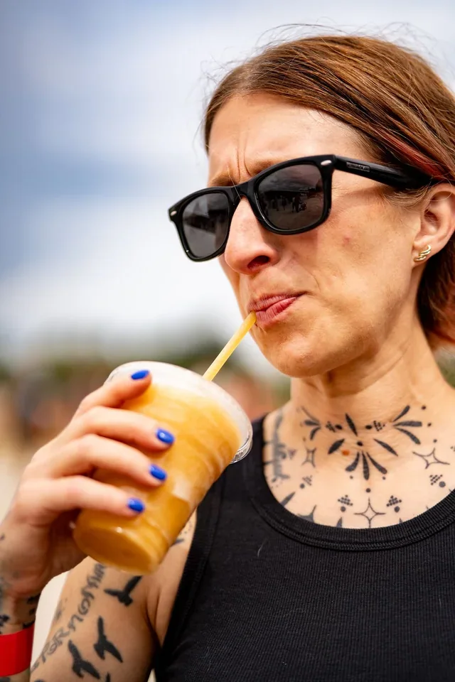 A tattooed person wearing sunglasses and sipping an iced drink outdoors